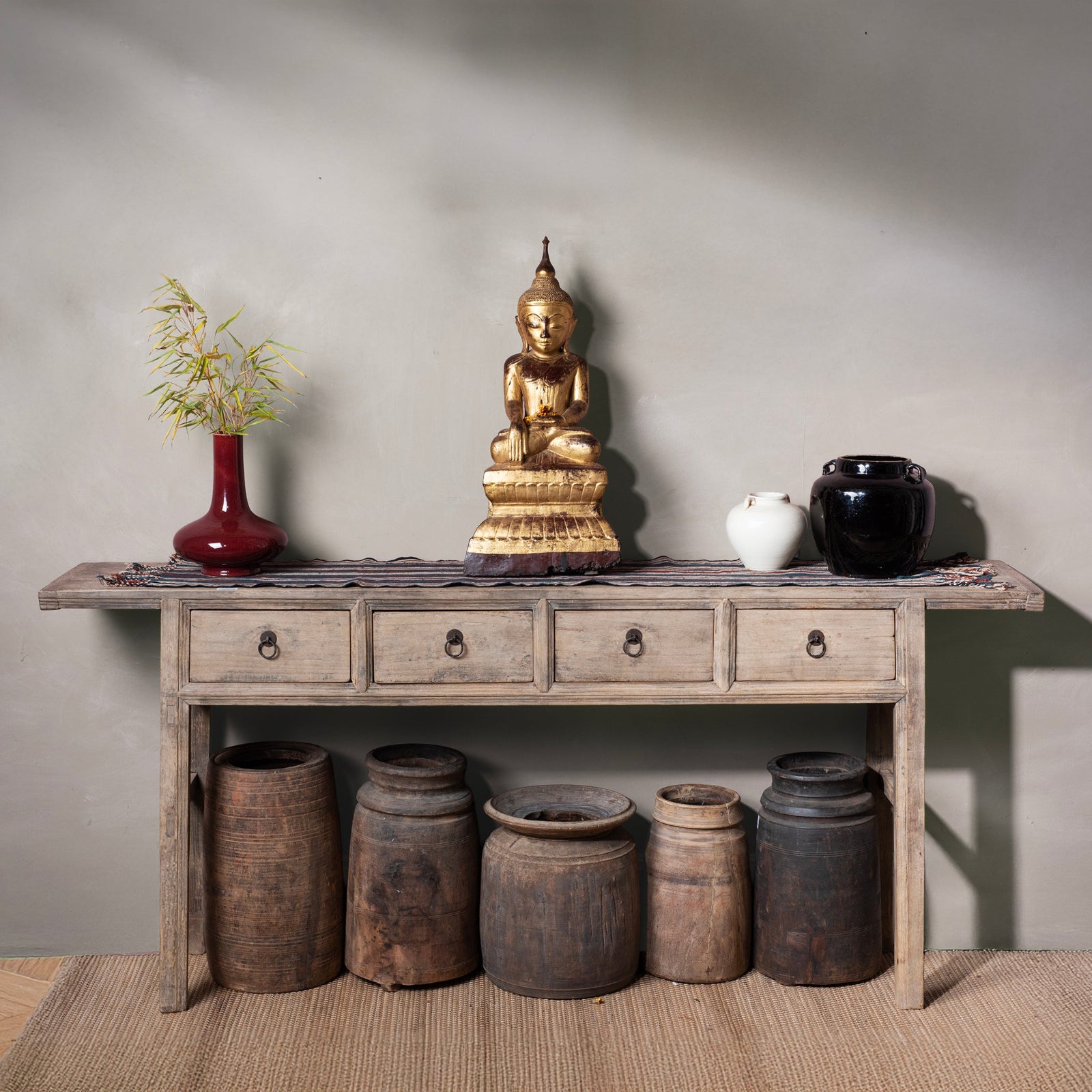 Wooden console table with decorative items including a gold Buddha statue, vases, and jars against a gray wall.