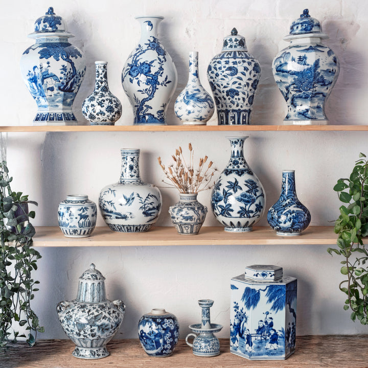 A collection of hand painted Chinese blue and white porcelain ceramic vases and canisters displayed on three wooden shelves against a white brick wall, with green plants framing the scene.