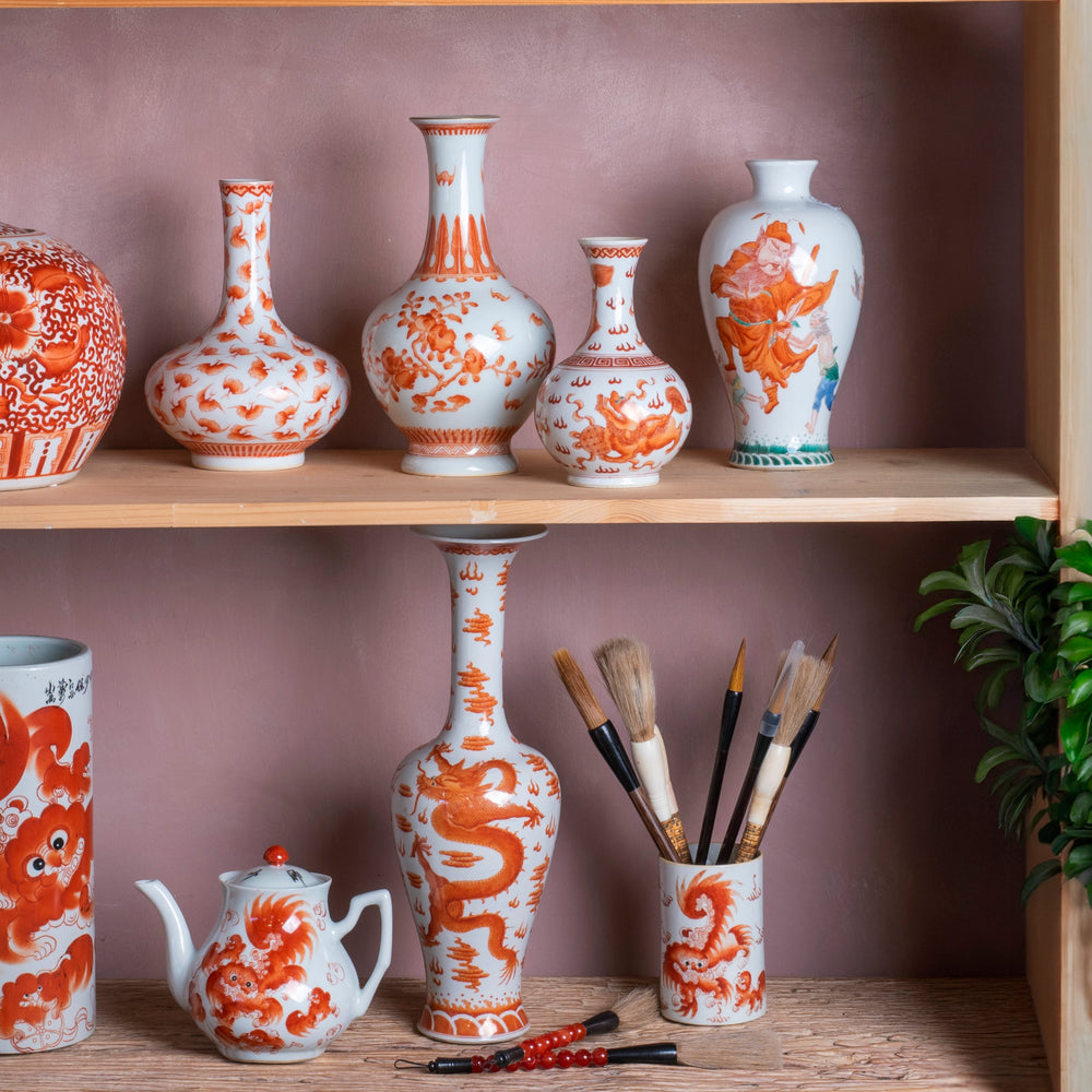 A collection of hand painted Chinese porcelain vases and vessels in a traditional orange and white style red oxide underglaze, displayed on wooden shelves against a dusty pink wall with green plants.