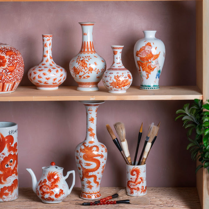 A collection of hand painted Chinese porcelain vases and vessels in a traditional orange and white style red oxide underglaze, displayed on wooden shelves against a dusty pink wall with green plants.